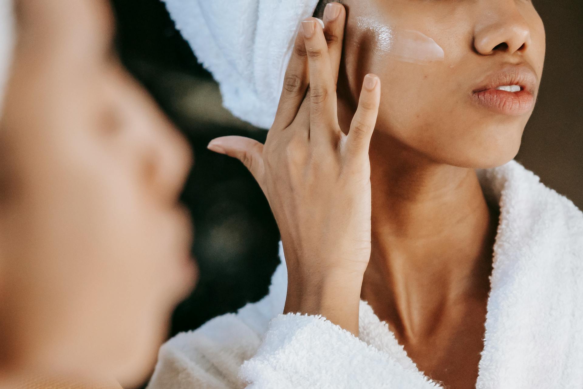 A woman in a bathrobe applying moisturizer on her face in front of a mirror.